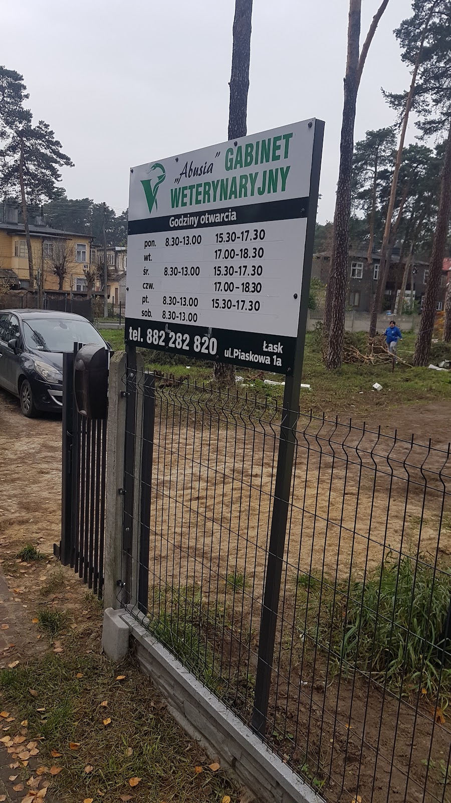 Biało-zielona tablica gabinetu Abusia z zielonym logo, godzinami otwarcia i numerem 882 282 820, w tle sosny i ogrodzenie.